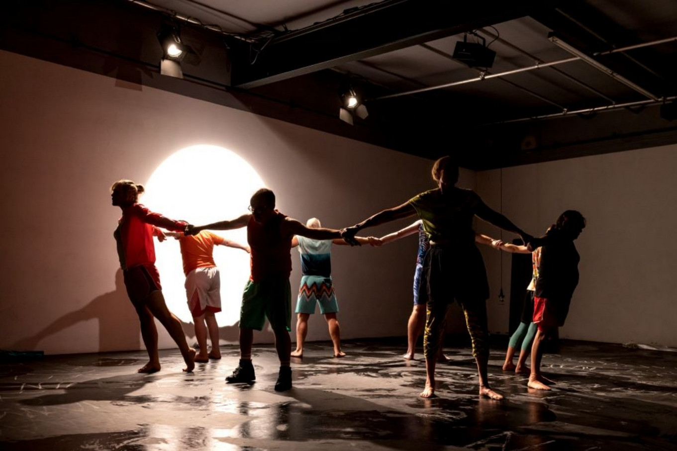 Eine Gruppe Menschen steht in einem wenig ausgeleuchteten Bühnenraum. Sie halten sich an den Händen und lehnen sich nach außen