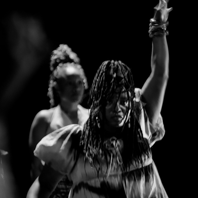 Schwarz weiß Bild. Zwei Personen stehen hintereinander in einem dunklen Raum. Die vordere Person hebt einen Arm