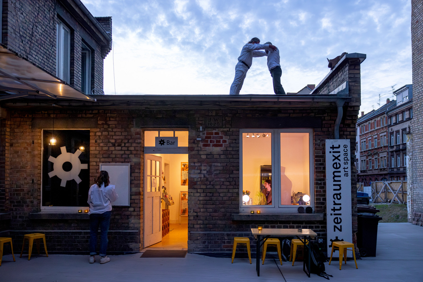 Eine Aussenansicht der Bar von zeitraumexit. Einem Backsteingebäude mit Flachdach. Auf dem Dach stehen zwei Personen und halten sich gegenseitig an den Armen