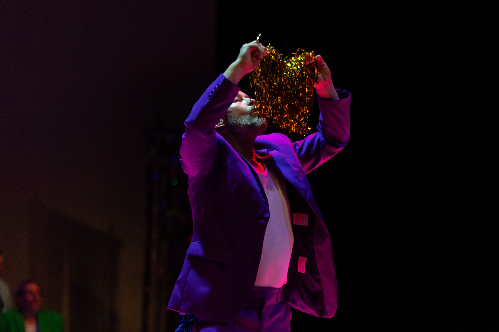 Person im lila Anzug steht in einem dunklen Raum und küsste einen goldenen PomPom