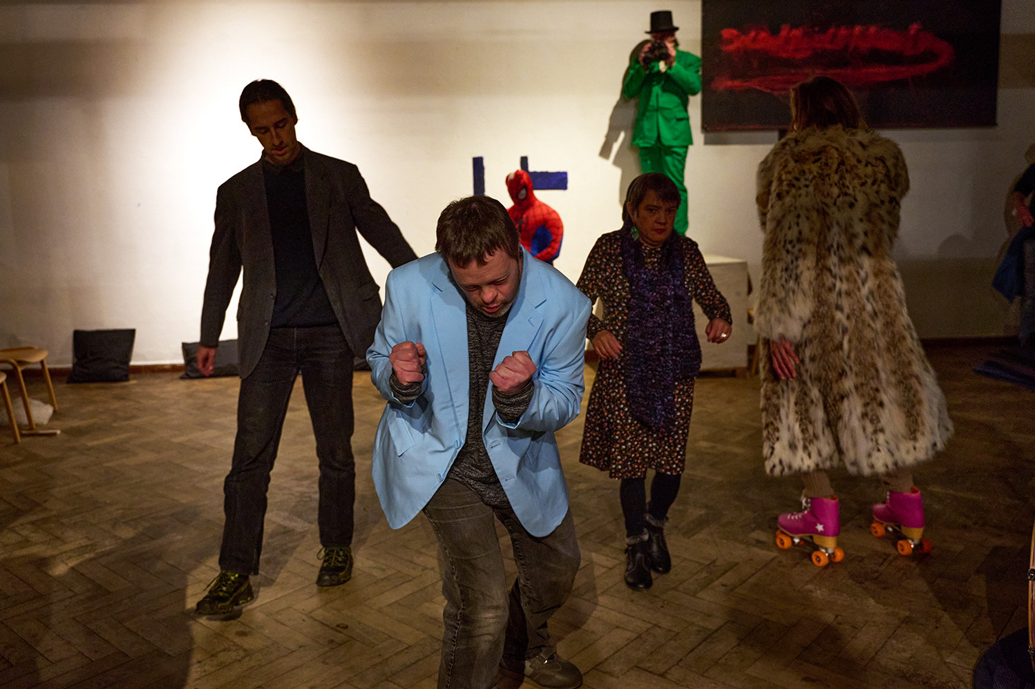 Vier Menschen bewegen sich im Hintergrund. Im Vordergrund steht eine Person im hellblauen Jackett mit geballten Fäusten