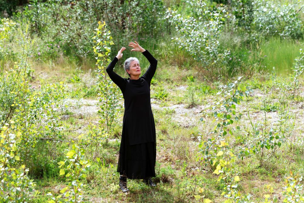 Eine Frau in einem schwarzen Kleid steht inmitten grüner Natur. Sie hält die Augen geschlossen und streckt die Arme nach oben