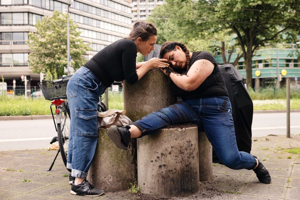 Zwei Frauen bewegen ihre Körper um einen Polter im Stadtraum. Die eine häuft Erde auf den Körper der Anderen