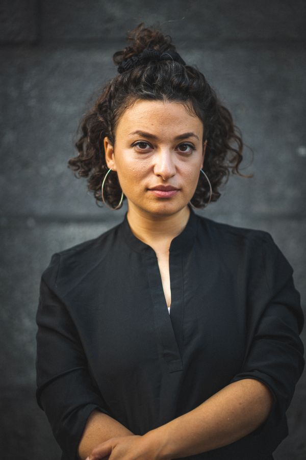 Porträtfoto von Laura Cazes. Vor grauem Hintergrund steht eine dunkelhaarige Frau in einem schwarzen Hemd und schaut in die Kamera.