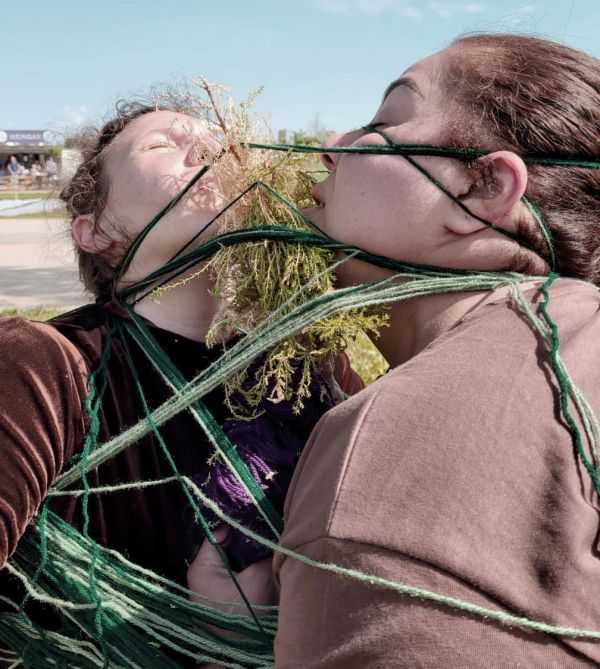 Uproot 2 - die Stadt Zwei Frauen stehen sich gegenüber. Sie sind mit Wollschnüren umwickelt. Zwischen ihnen Grünzeug