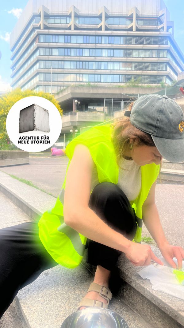Utopisches Werkeln Eine Frau in gelber Warnweste sitzt vor einem brutalistischen Gebäude auf einer Treppe. Sie schreibt etwas.