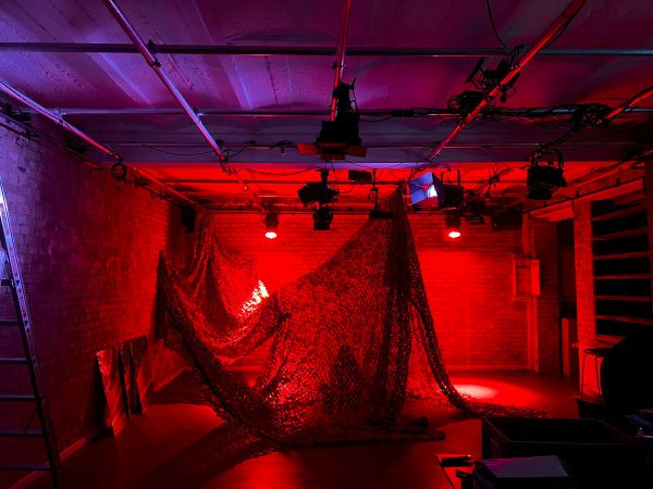 Camouflage Tarnnetz mit verschiedenen schemenhaften Personen in einem dunklen, rot beleuchteten Theaterraum