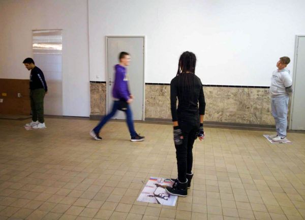 Gesicht I Gesicht Jugendliche befinden sich in einem weißen Raum, mit beige gekacheltem Fußboden. Eine Person mit langen Haaren im Vordergrund steht im Vordergrund. Die Jugendlichen im Hintergrund gehen hin und her.
