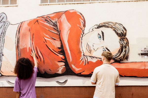 Yollar Eine Person mit langem Haar und eine Person mit kurzem Haar stehen mit dem Rücken zur Kamera und berühren das Graffiti einer liegenden Frau an der Wand