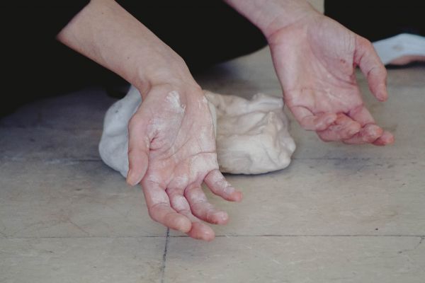 Körper, Spuren, Spüren Zwei Hände zeigen mit den Handflächen nach oben in die Kamera. Sie sind mit weißem Ton verschmiert. Im Hintergrund liegt ein Tonklumpen auf dem Boden.
