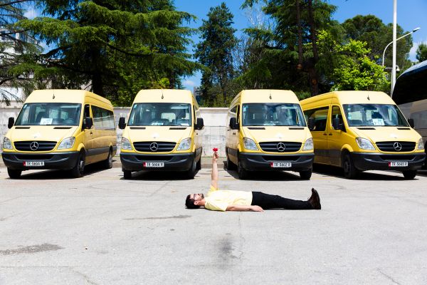 Performance Art Studies Mann in gelbem T-Shirt und schwarzer Hose liegt vor gelb-schwarzen Vans auf der Straße und streckt einen Arm mit roter Blume in die Luft