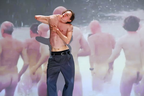 Making of a Man Eine Person steht vor einer Videoleinwand, auf der nackte Menschen ins Wasser laufen. Die Person zieht sich einen Gummioberkörper aus