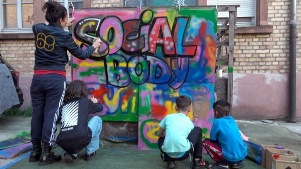 Varieté Utopolis Vier Kinder sitzen und stehen vor einem Plakat, auf das sie ein buntes Graffiti malen und sprayen