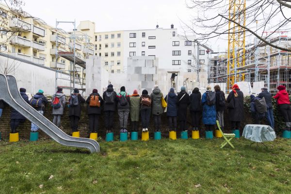 Mobile Albania Viele Menschen stehen aufgereiht auf Eimern auf einer grünen Wiese mit dem Rücken zum Betrachter. Vor ihnen steht eine eiserne Rutsche und ein runder Tisch mit karierter Tischdecke. Die Menschen schauen über eine Mauer in eine Baustelle.