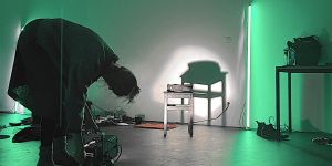 Das Foto zeigt den Schemen einer Frau, die einen Scheinwerfer auf einen Hocker vor einer weißen Wand richtet. In der Ecke lehnt eine grün leuchtende Neonröhre. 