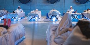 Foto eines steril weiß beleuchteten Raumes mit Feldbetten, in denen Menschen liegen. Das Foto ist aus der Perspektive eines Liegenden aufgenommen und zeigt dessen Füße und die belegten Betten an der gegenüberliegenden Wand