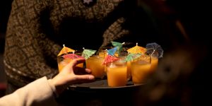 Eine Hand greift nach einem orangenen Drink mit Schirmchen auf einem Tablett